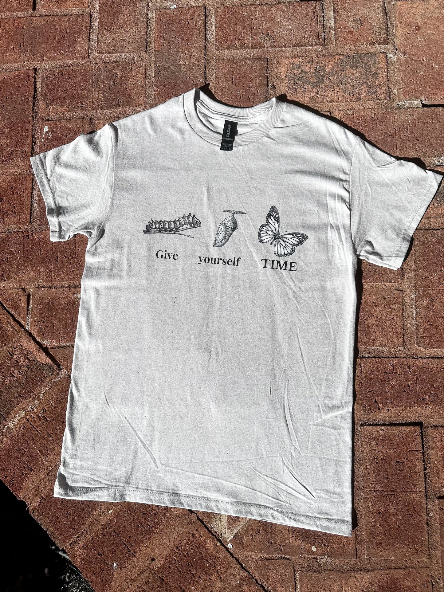 Short sleeve ice grey butterfly graphic t-shirt with self-love quote, flat lay on brick surface