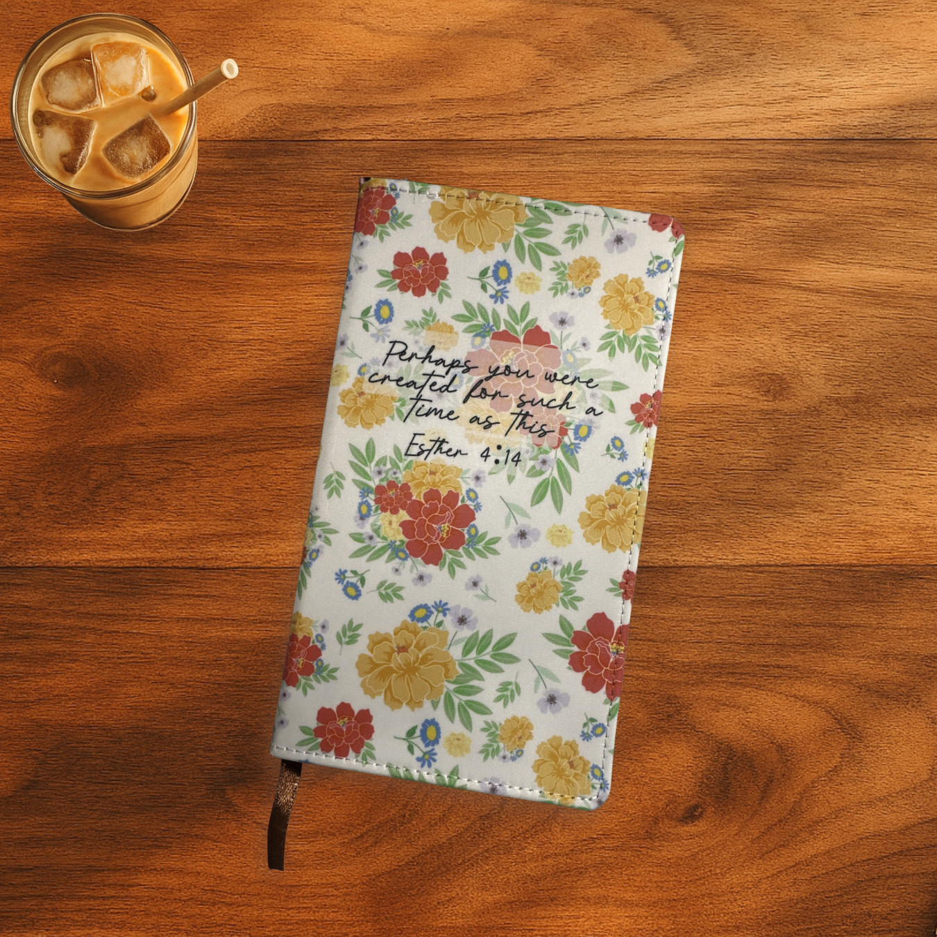 Floral polyurethane notebook with Esther 4:14 verse on the front, the notebook is on a wooden surface next to a glass of iced coffee.