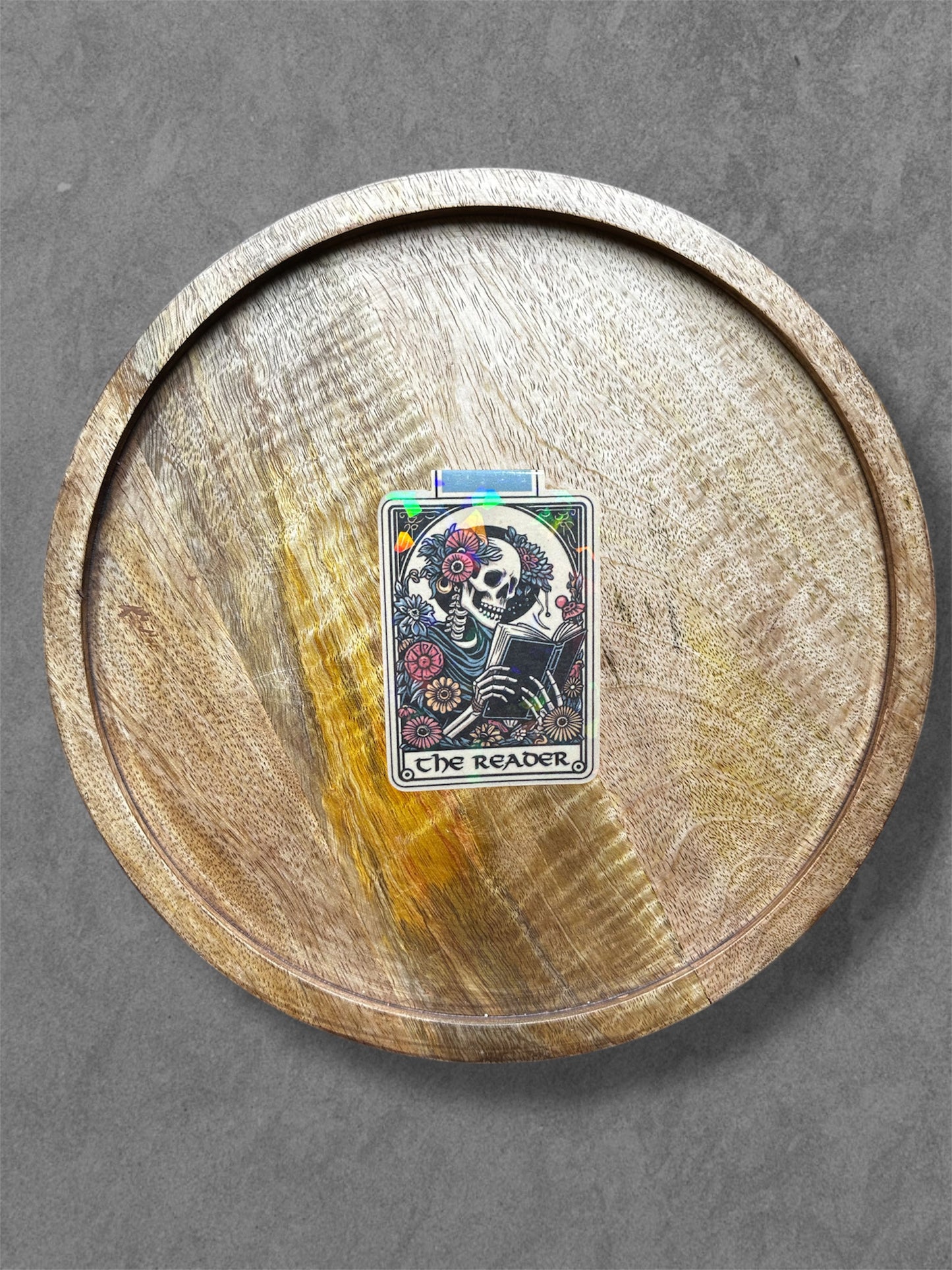 Wooden circular tray with a tarot card design featuring a skeleton reading and flowers magnetic bookmark on a gray background.