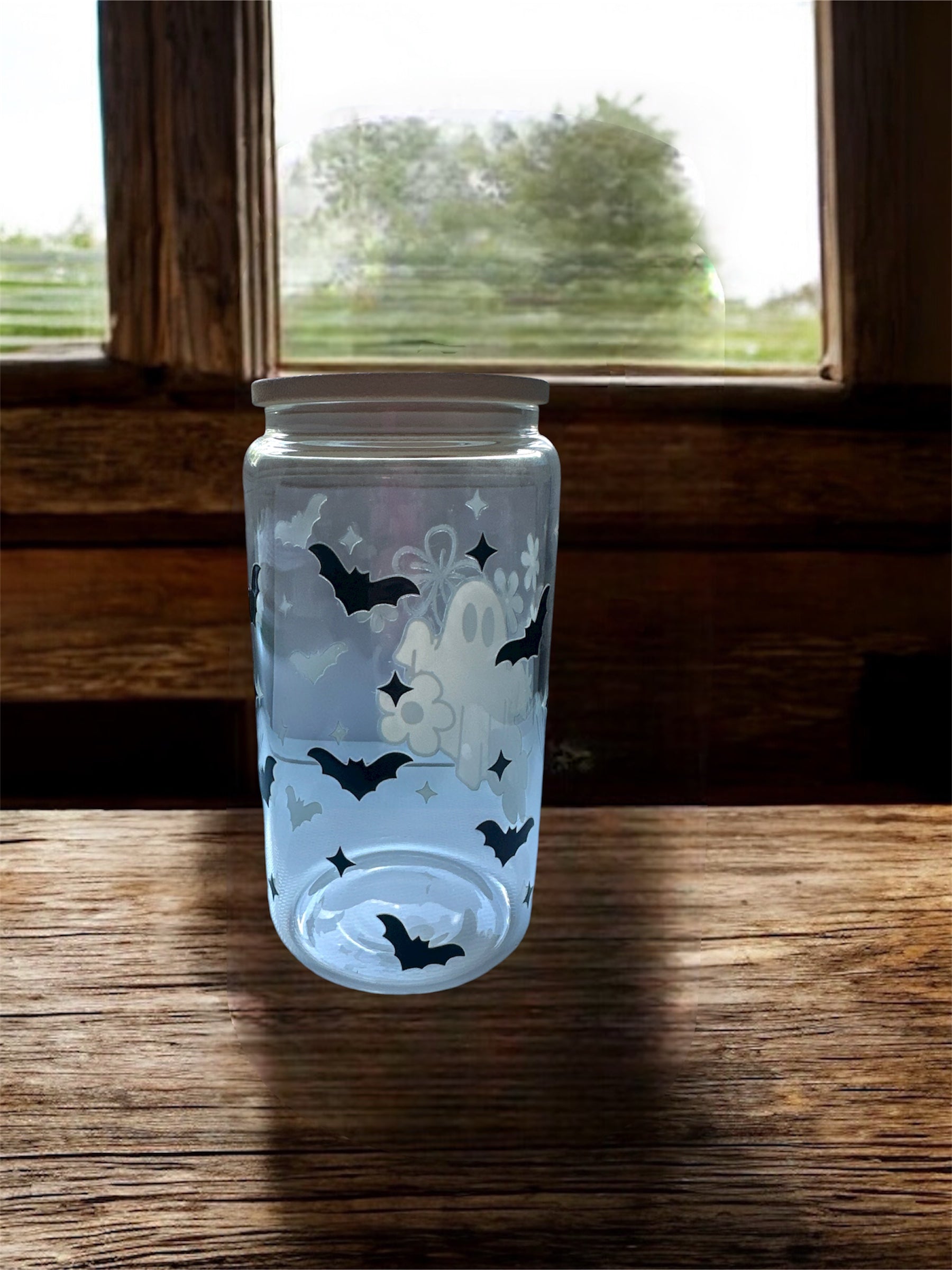 Clear 16 oz glass tumbler with wooden lid. Tumbler has a black graphics of  flowers and sparkles. Tumbler is sitting on a wooden surface.