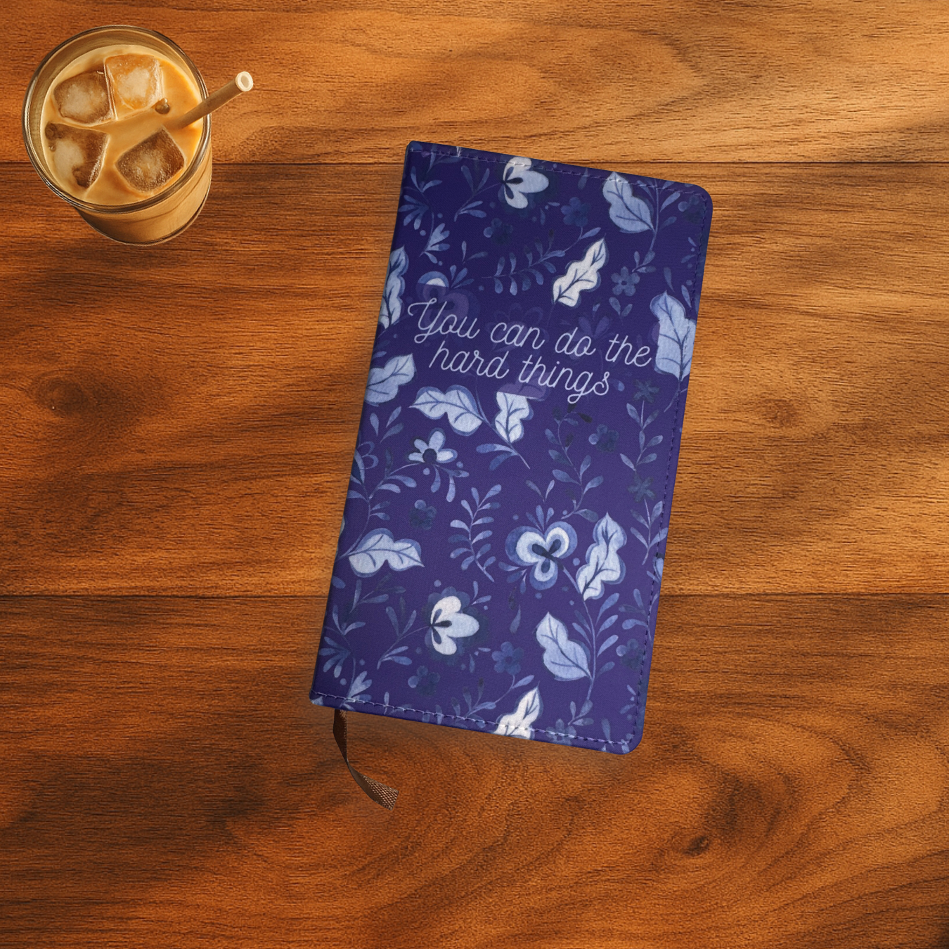 Blue floral polyurethane notebook with 'You can do the hard things' on the front, the notebook is on a wooden surface next to a glass of iced coffee.