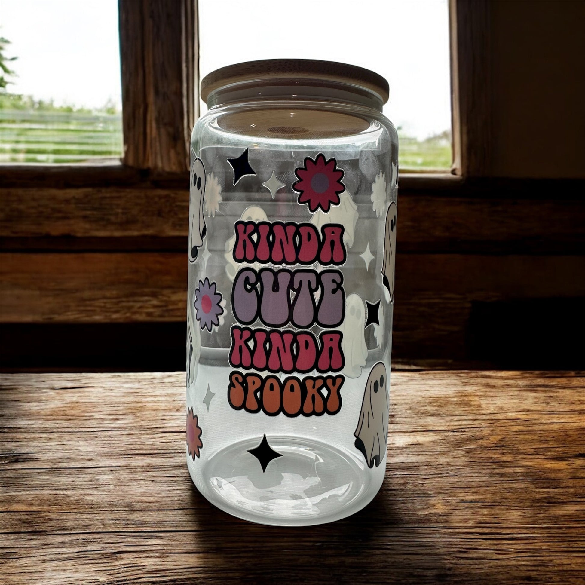 Clear 16 oz glass tumbler with wooden lid that says colorful 'Kinda Cute Kinda Spooky' with ghost, flowers, and sparkle graphics. Tumbler is sitting on a wooden surface.