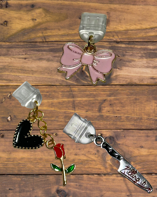 Three decorative dust plugs on a wooden surface: a pink bow, a black heart with a red rose, and a silver knife.