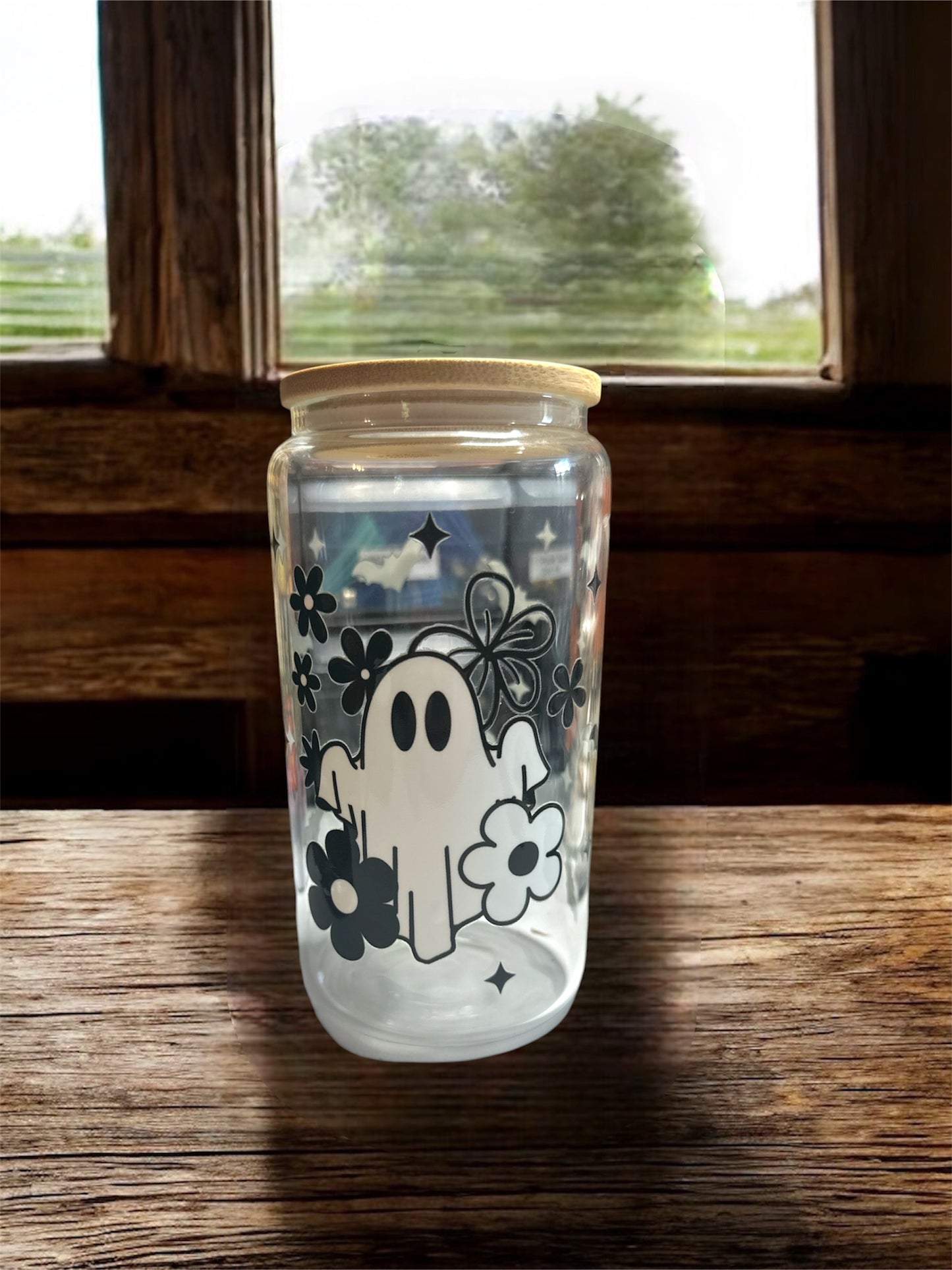 Clear 16 oz glass tumbler with wooden lid. Tumbler has a black and white ghosts with flowers and sparkles graphics. Tumbler is sitting on a wooden surface.