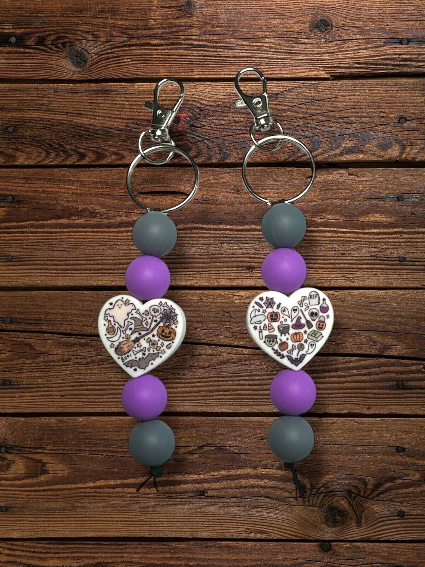 Two white heart-shaped silicone beaded keychains with Halloween-themed items inside, with purple and gray beads attached to silver hardware keychains, on a wooden surface