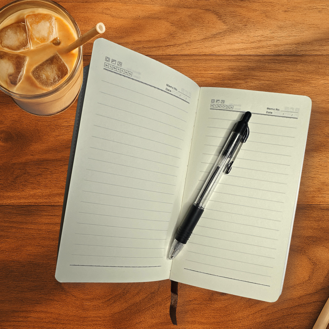 Open notebook with a pen on a wooden surface next to a glass of iced coffee.