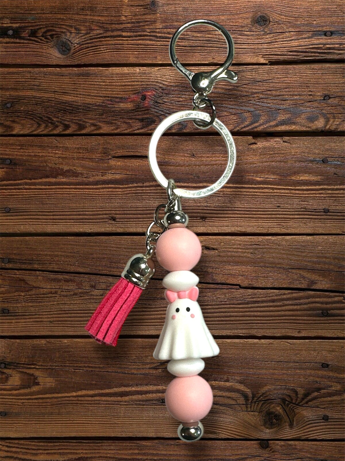 silicone white ghost with pink bow and blush on a silver keychain hardware with pink and white silicone beads and a pink fringe tassel on silver keychain hardware on a wooden table
