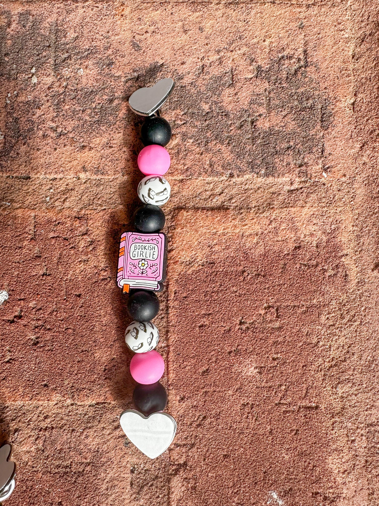 back of Silicone beaded Kindle/ e-reader strap with pink ‘bookish girlie’ book focal bead and pink, black, white, and book-patterned beads, shown on a brick background.