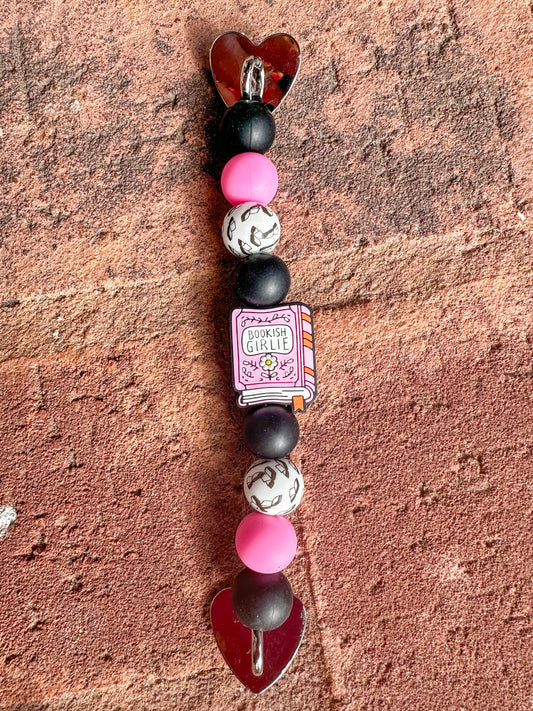 close up of one Silicone beaded Kindle/ e-reader strap with pink ‘bookish girlie’ book focal bead and pink, black, white, and book-patterned beads, shown on a brick background.