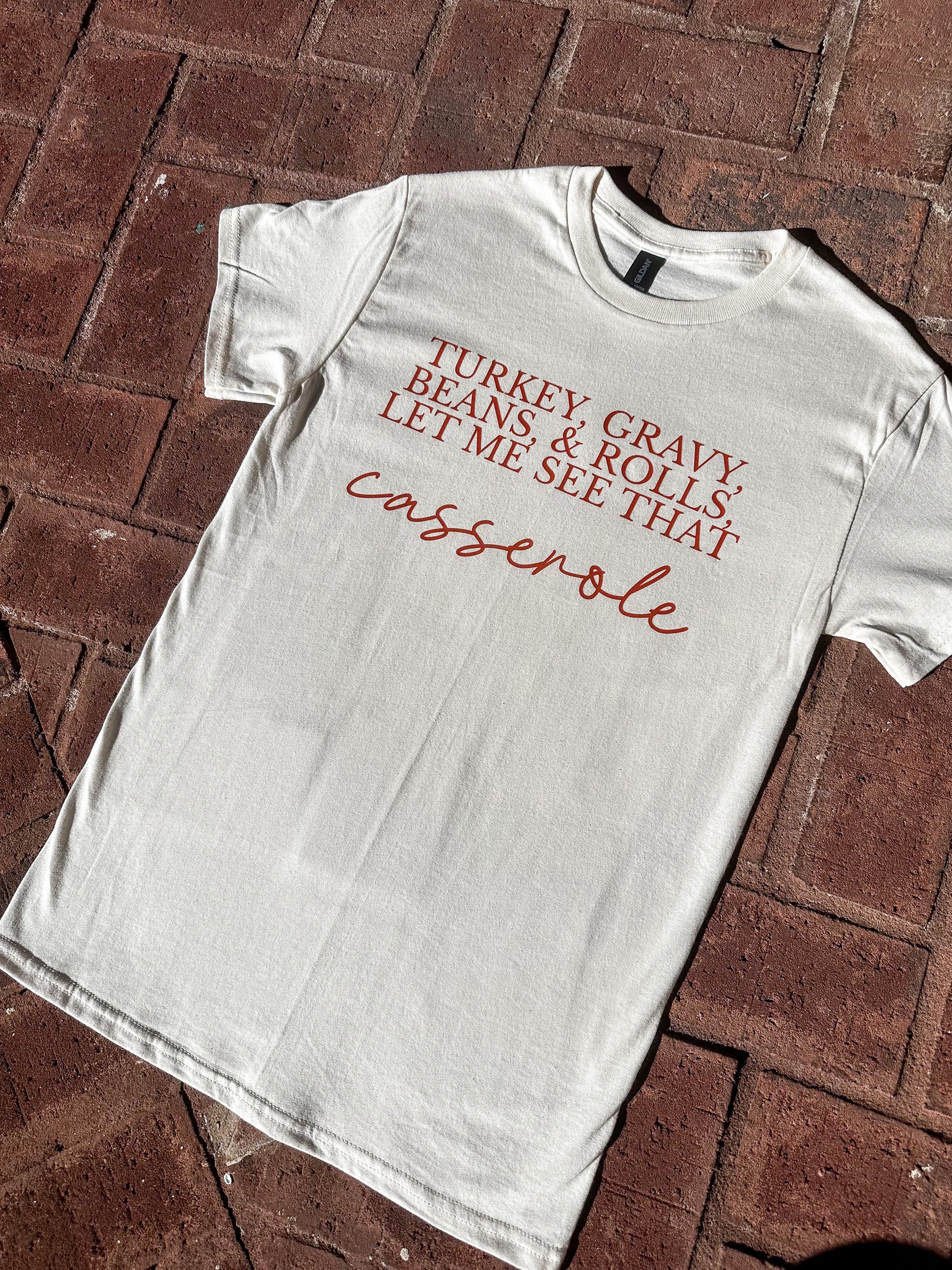 Funny Thanksgiving graphic t-shirt on natural Gildan G500 with ‘Turkey, Gravy, Beans & Rolls’ design, flat lay on brick floor.