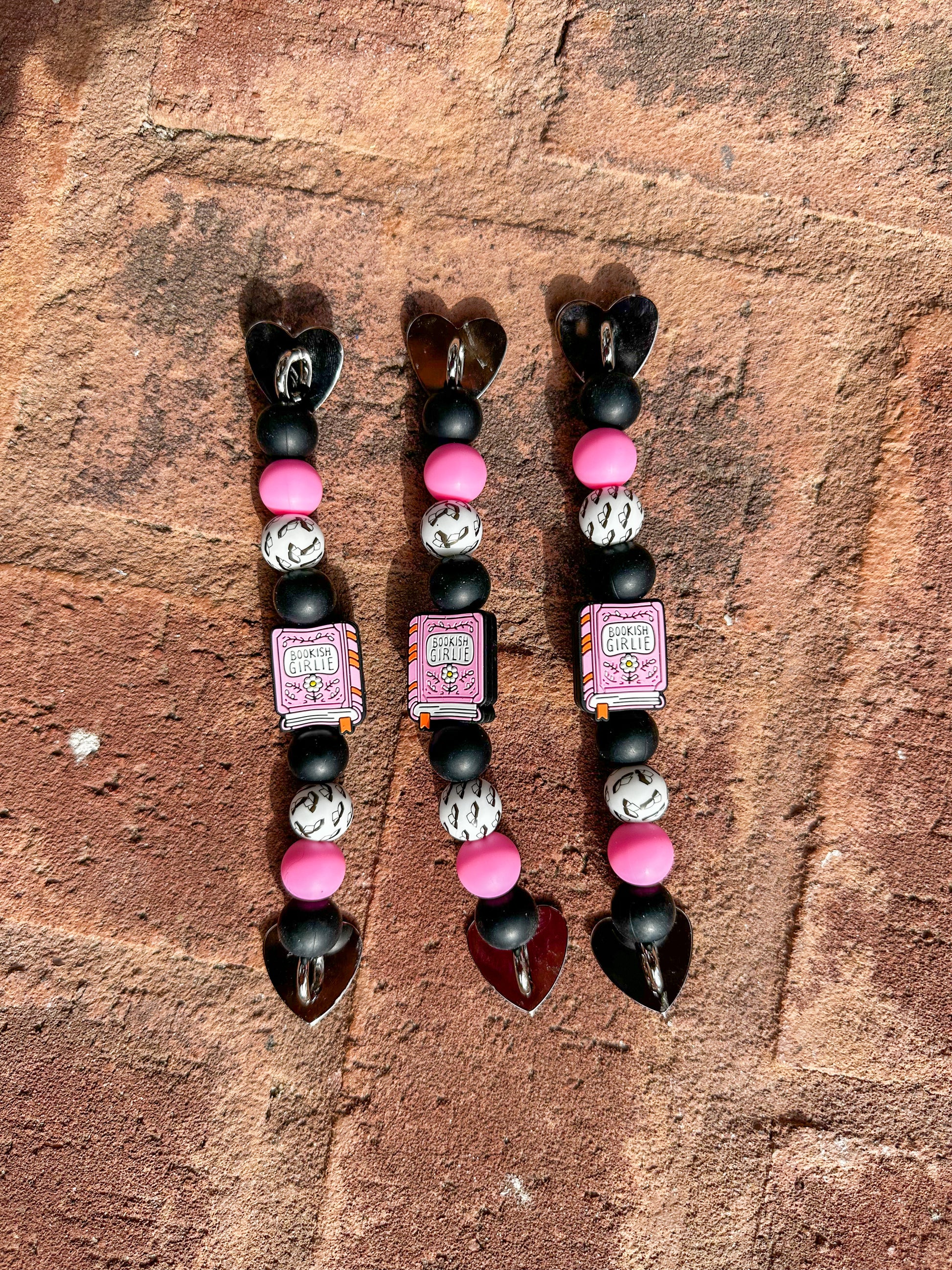 3 Silicone beaded Kindle/ e-reader straps with pink ‘bookish girlie’ book focal bead and pink, black, white, and book-patterned beads, shown on a brick background.