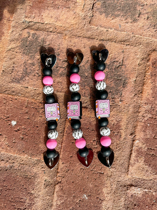 3 Silicone beaded Kindle/ e-reader straps with pink ‘bookish girlie’ book focal bead and pink, black, white, and book-patterned beads, shown on a brick background.
