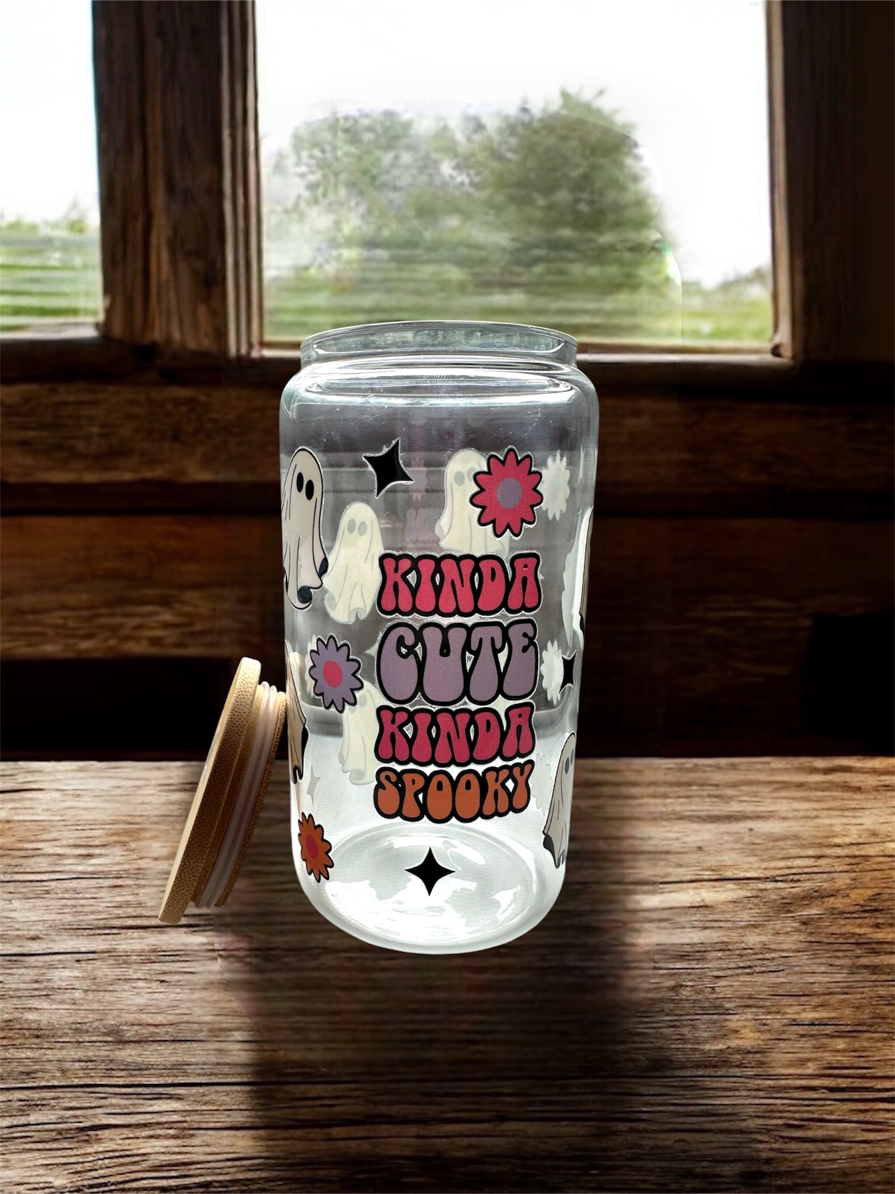 Clear 16 oz glass tumbler with wooden lid taken off. Glass says colorful 'Kinda Cute Kinda Spooky' with ghost, flowers, and sparkle graphics. Tumbler is sitting on a wooden surface.