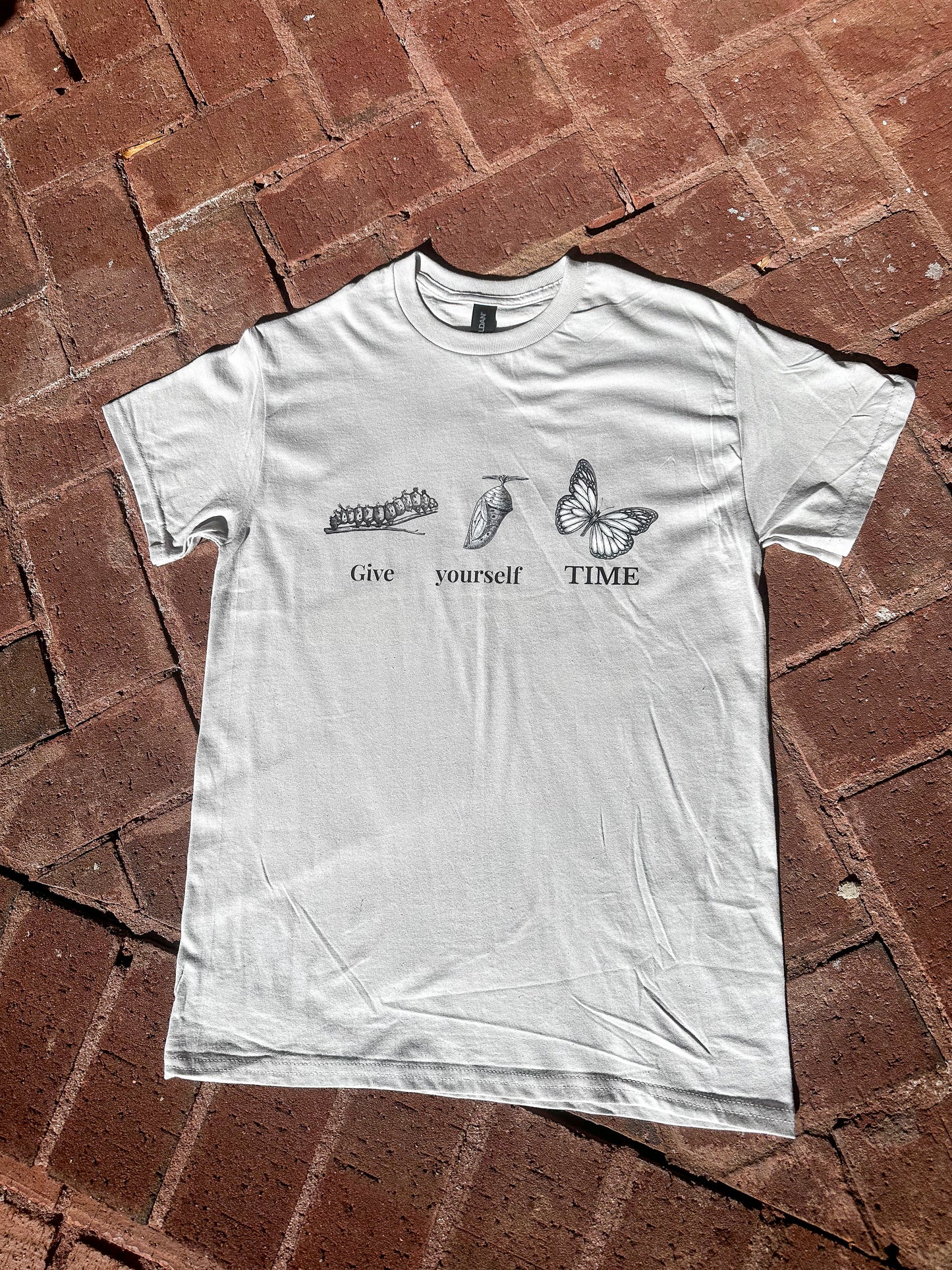 Vintage butterfly transformation shirt in ice grey laid flat on brick background.