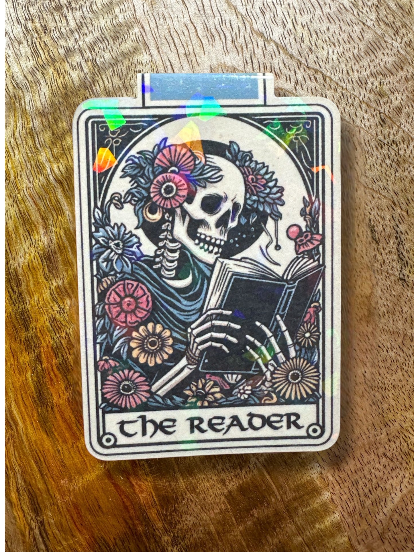 Holographic magnetic bookmark of a skeleton reading a book with floral elements on a wooden surface