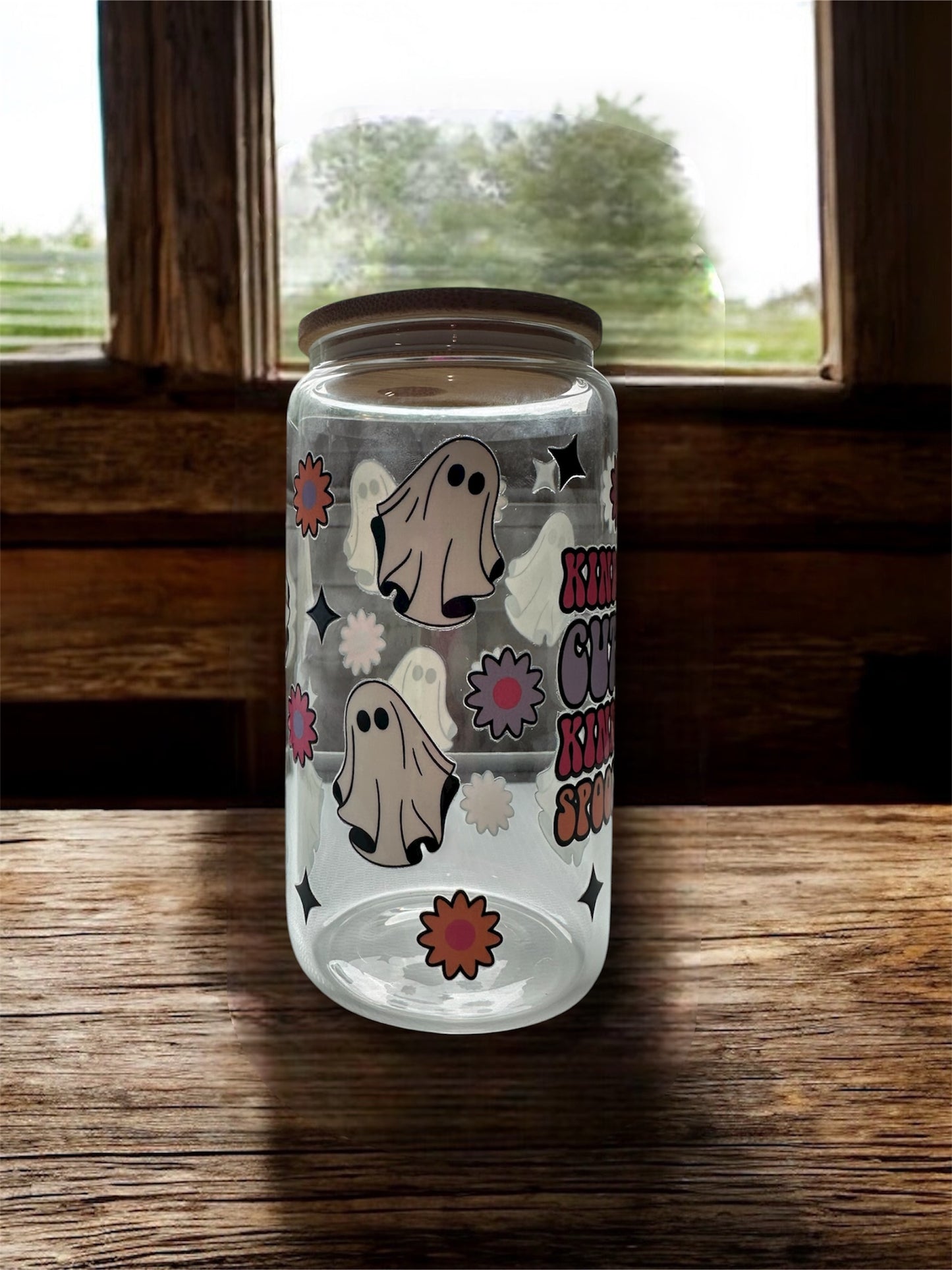 Clear 16 oz glass tumbler with wooden lid that says colorful 'Kinda Cute Kinda Spooky' with ghost, flowers, and sparkle graphics. Tumbler is sitting on a wooden surface.